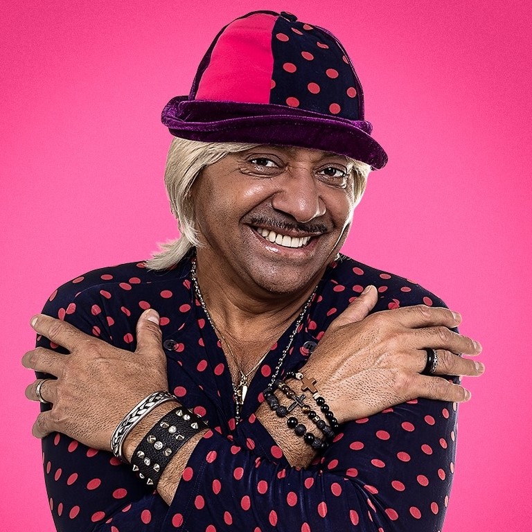 Homem sorrindo com chapéu e roupa estampada sobre fundo rosa